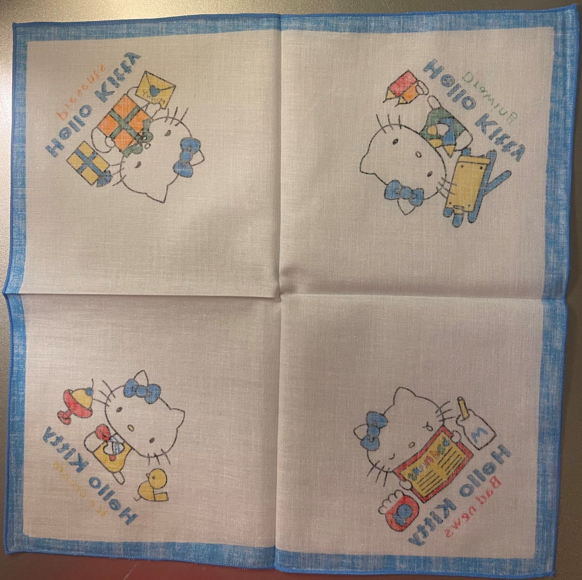 Unfolded cotton napkin with blue border showing four Hello Kitty illustrations in corners with colored kites, toys, and Japan