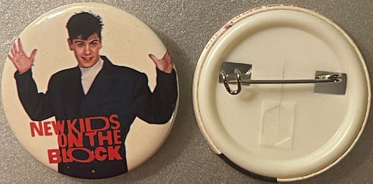 Front and back of a round button pin featuring a young man in a black jacket with raised hands and red text reading "New Kids