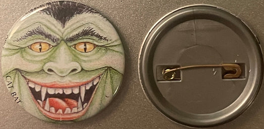 Vintage pinback button with grotesque green vampire face with fanged grin and yellow cat eyes, shown front and back against g