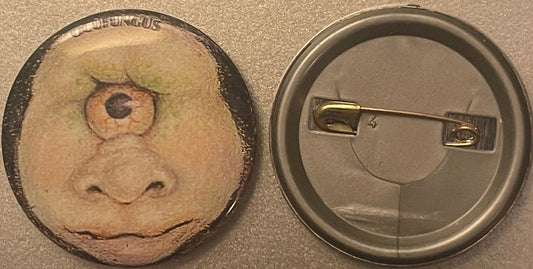 Front and back view of a tan pinback button featuring an embossed one-eyed cyclops face with green hair and brown eye, displa