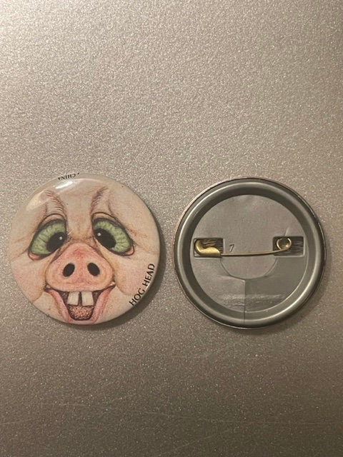 Front and back of a round pinback button featuring a grotesque pig face with green eyes, pink snout, and buck teeth on beige
