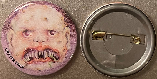 Vintage pinback button with grotesque face featuring purple-tinted skin, closed eyes, and prominent teeth, shown front and ba