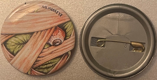 Front and back of a round pin badge featuring a mummified face wrapped in bandages with the word "MUMMEYE" printed at top, sh