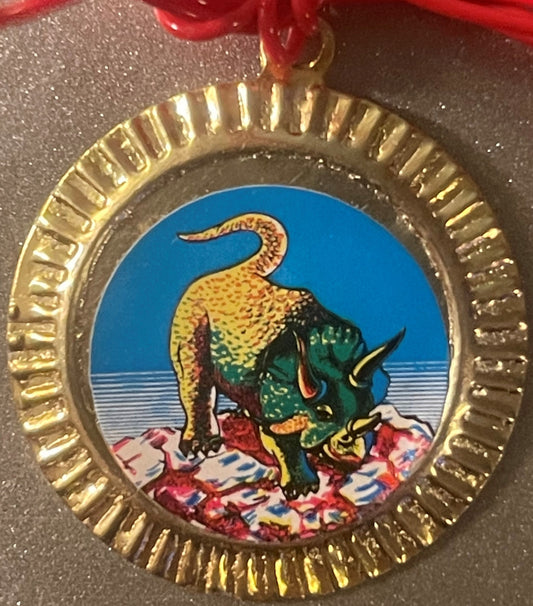 Circular brass charm with scalloped edge featuring colorful dinosaur illustration: yellow spotted creature with green body an