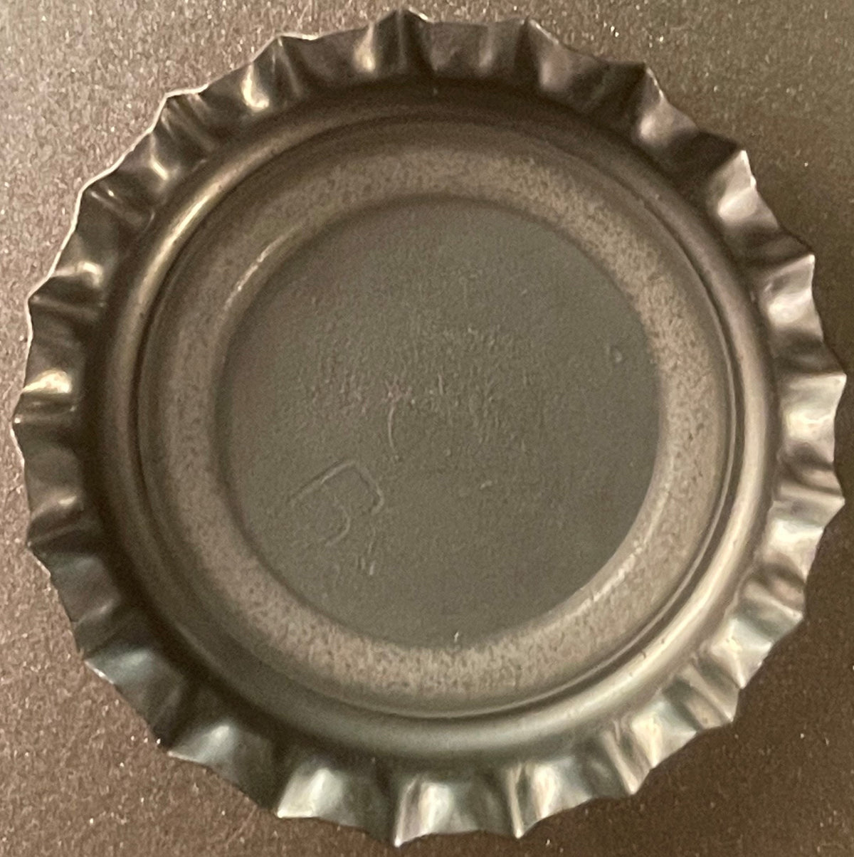 Vintage metal bottle cap with crimped fluted edges, viewed from above on gray surface, showing embossed circular center detai