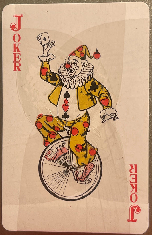 Joker playing card from vintage deck featuring colorful clown on unicycle holding spade card, yellow suit with red pompoms, a