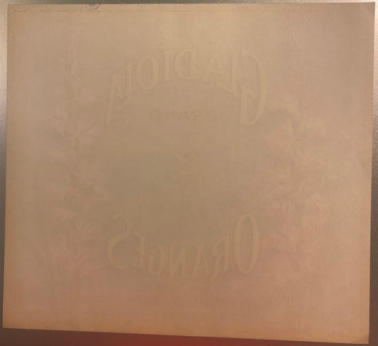 Faded embossed fruit crate label with "Gladiola" and "Covina" text visible on aged tan cardboard, photographed flat against d