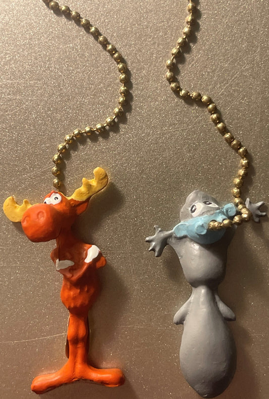 Two vintage character fan pull chains suspended from gold beaded strings against a textured gray wall: an orange moose with y
