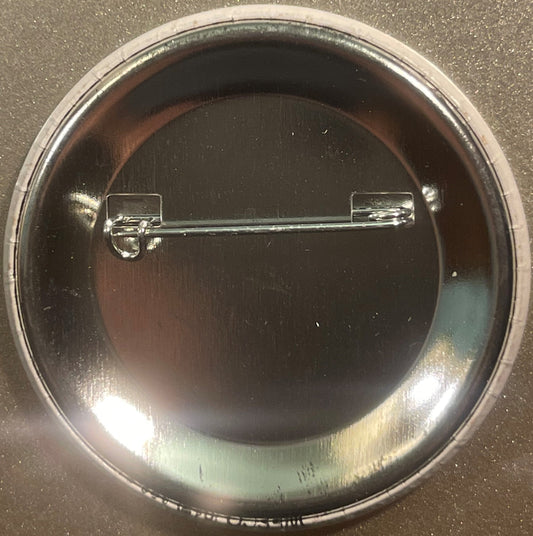 Overhead view of a circular metal pin badge with chrome safety pin backing, dark face, displayed on textured surface