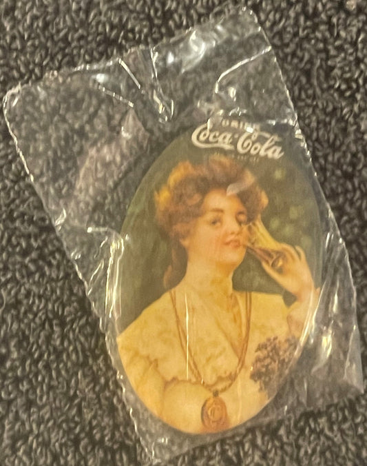 Unopened plastic-wrapped vintage Coca-Cola mirror featuring a woman in yellow dress, printed "Coca-Cola" text visible at top,
