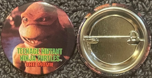 Front and back of a circular pin featuring Michelangelo from Teenage Mutant Ninja Turtles movie, with green text logo on oran
