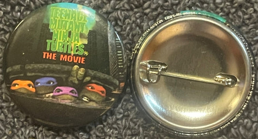 Vintage TMNT movie pin showing four colored turtle masks (orange, blue, purple, red) with green sewer backdrop, displayed fro