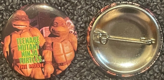 Vintage TMNT movie pin with orange turtle characters and green text on front; back shows silver metal pinback clasp on textur