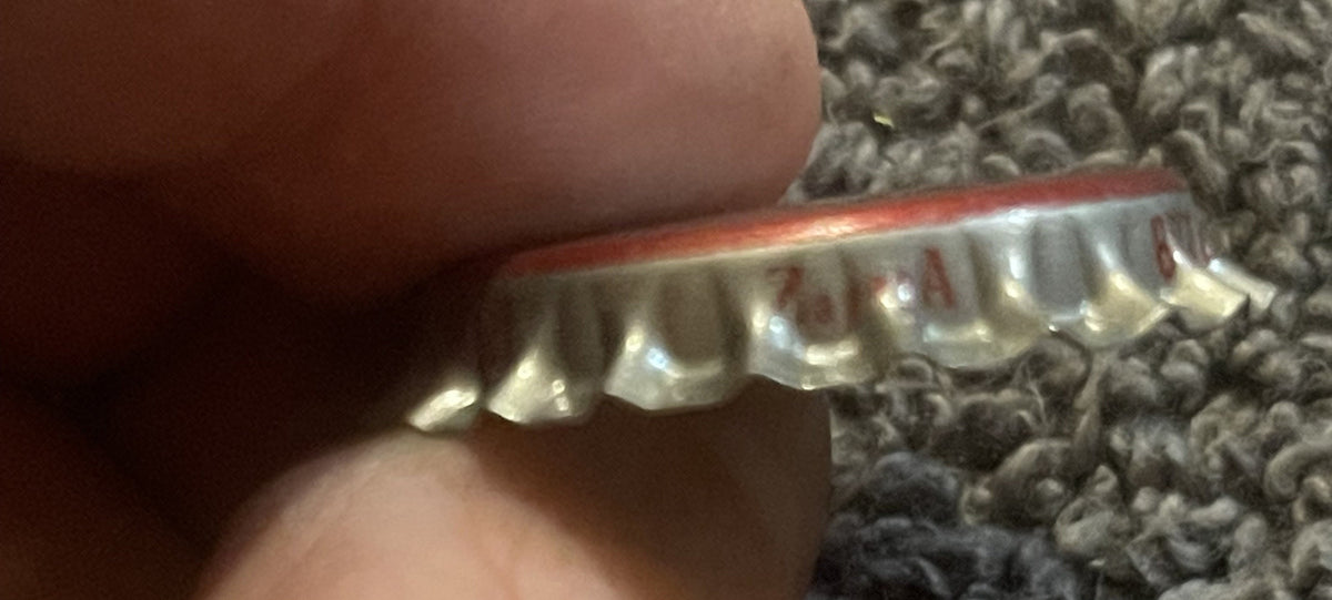 Vintage bottle cap with red rim and white ridged underside held by a finger against textured gray surface