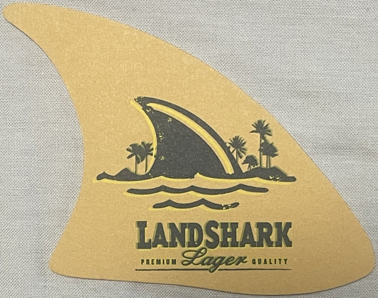 Tan shark fin-shaped coaster with black silhouette of shark fin, palm trees, and waves, printed text reading "LANDSHARK" and