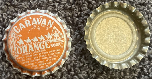 Two vintage bottle caps on textured fabric: orange Caravan Orange Soda cap with mountain design, and plain metallic cap shown
