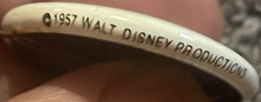 Close-up of cream-colored pinback button rim embossed with "©1957 WALT DISNEY PRODUCTION," held by a person's finger for scal