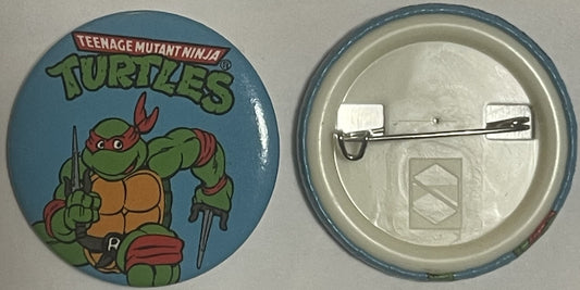 Front and back of a blue circular TMNT pinback button showing Raphael with sai and sword, metal pin fastener visible on rever