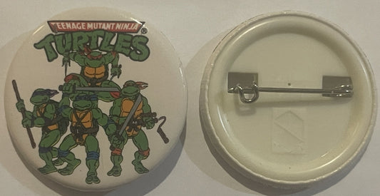 Front and back of round white button pin featuring four green cartoon turtles in battle poses with "Teenage Mutant Ninja Turt
