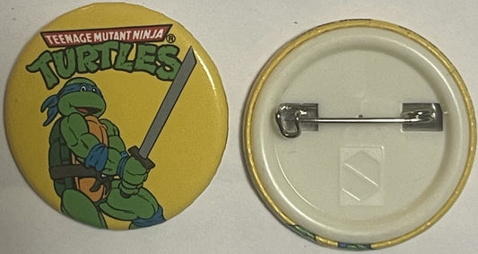 Front and back of a yellow circular pin featuring Leonardo from Teenage Mutant Ninja Turtles with blue bandana holding a kata
