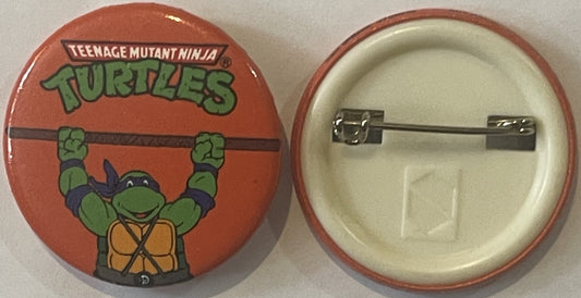 Front and back of a red circular pin featuring a green turtle character with "Teenage Mutant Ninja Turtles" text, shown with
