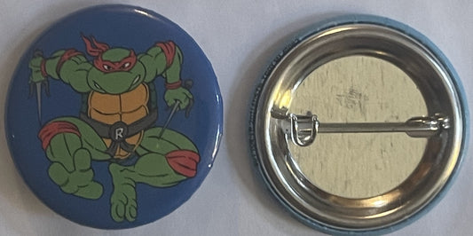 Front and back of a blue pinback button featuring a green Teenage Mutant Ninja Turtle with red mask and "R" on chest, shown a