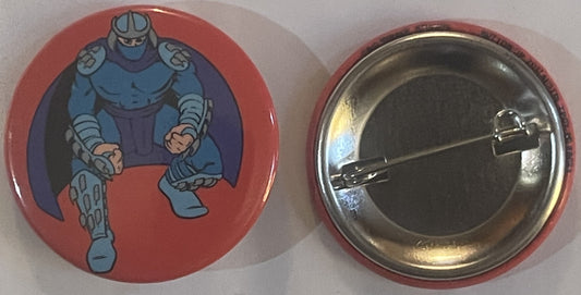 Front and back of a red circular pin featuring Shredder from TMNT in blue armor on coral background, with metal clasp visible