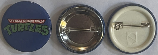 Three views of a round pin-back button: front showing blue background with green "Teenage Mutant Ninja Turtles" text, back sh
