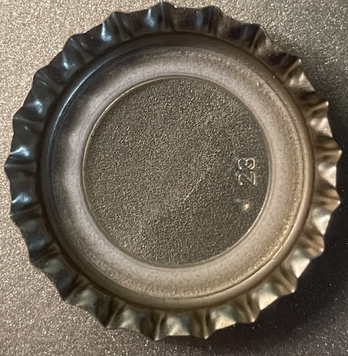 Overhead view of a ridged metal bottle cap with textured center surface and small maker's mark visible, on gray background