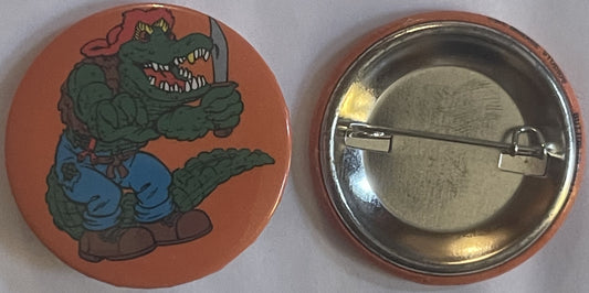 Front and back of a round orange pin featuring a green cartoon alligator character in blue pants holding a sword against a re