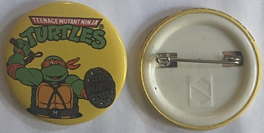 Front and back of yellow TMNT Michelangelo pin showing cartoon turtle character with nunchaku and metal pin fastener on rever