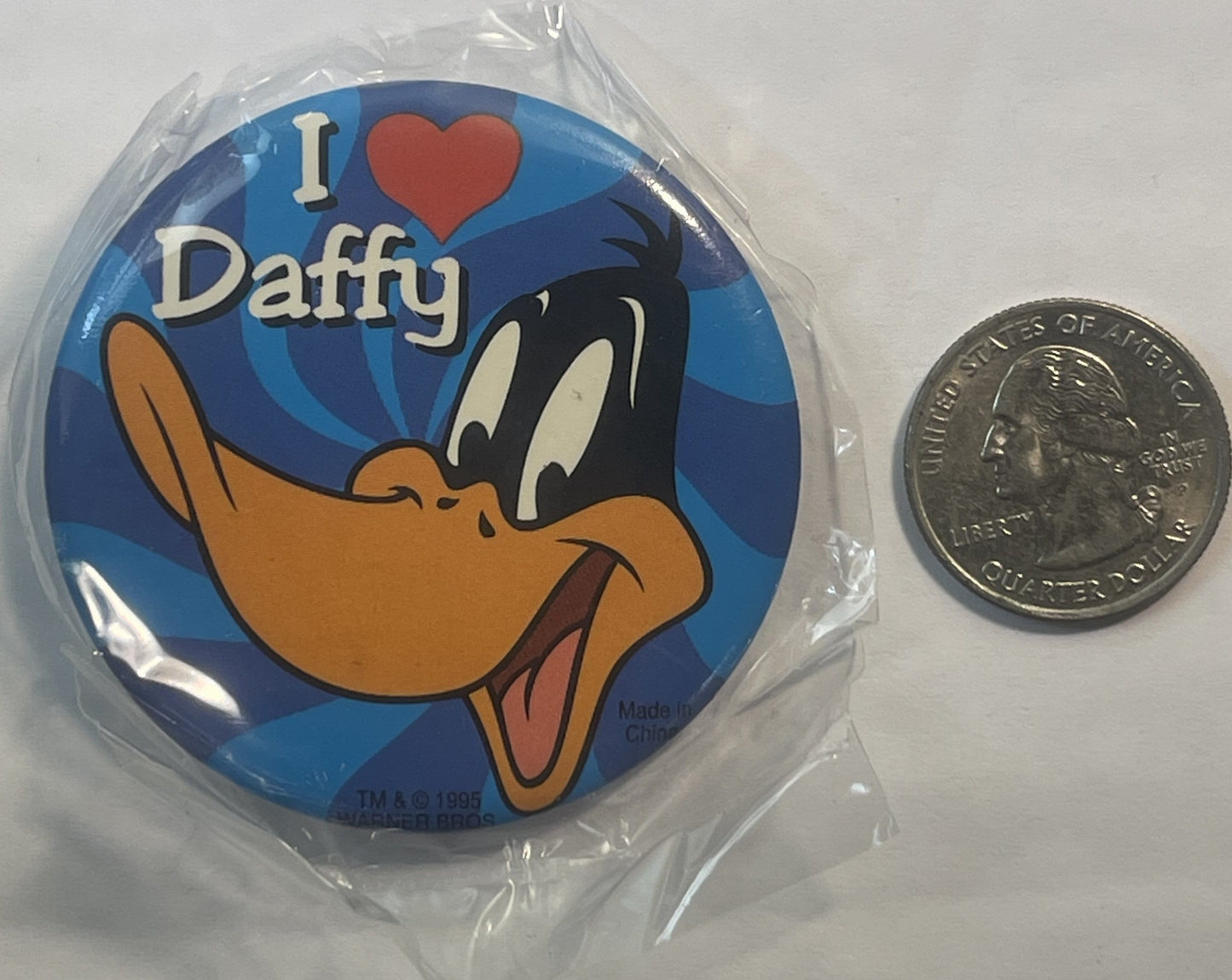 Unopened 1995 Daffy Duck pin in plastic wrapper next to a quarter for scale, showing cartoon duck with "I ♥ Daffy" text on bl