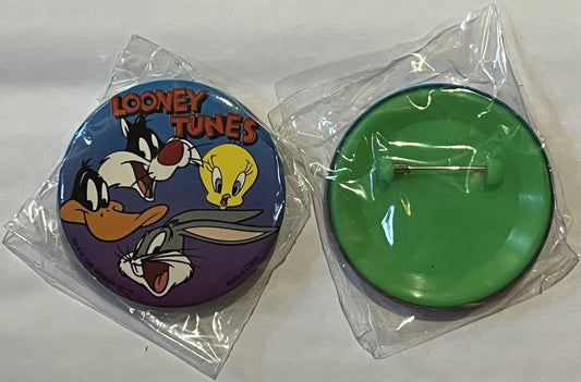 Two unopened circular pins in clear plastic packaging: left shows Looney Tunes characters on blue background, right is solid