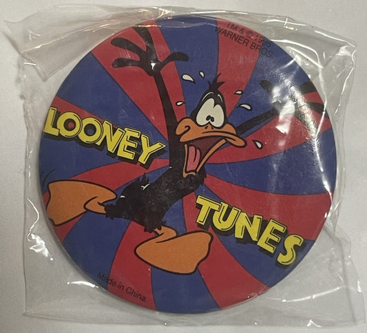 Circular pin featuring Daffy Duck with "LOONEY TUNES" text on red and blue radiating background, sealed in clear plastic pack