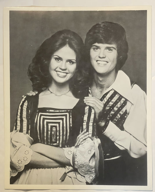 Black and white photograph of a young man and woman in 1970s-style clothing holding decorative fans, smiling at the camera
