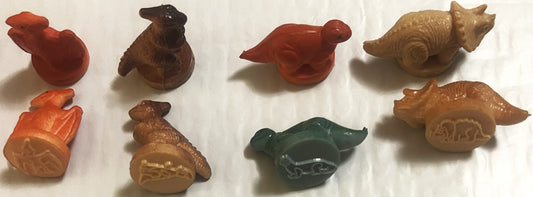 Eight small colored rubber dinosaur and animal stamps arranged on white surface, showing red, brown, tan, and green vintage t