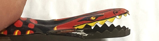 Vintage red and black snake tin clicker toy with open mouth showing yellow teeth, held in a hand for scale.