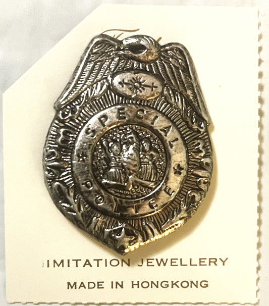 Silver-toned police badge pin with eagle on cream cardboard backing labeled "IMITATION JEWELLERY MADE IN HONGKONG