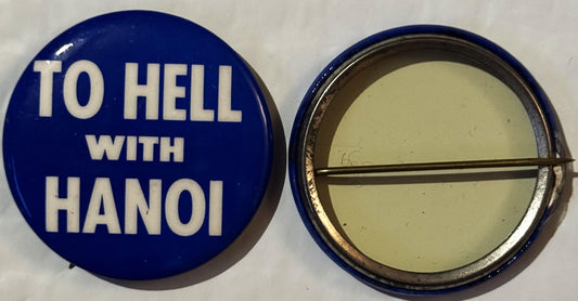 Front and back of a blue pinback button reading "TO HELL WITH HANOI" in white capital letters, with metal clasp visible on re