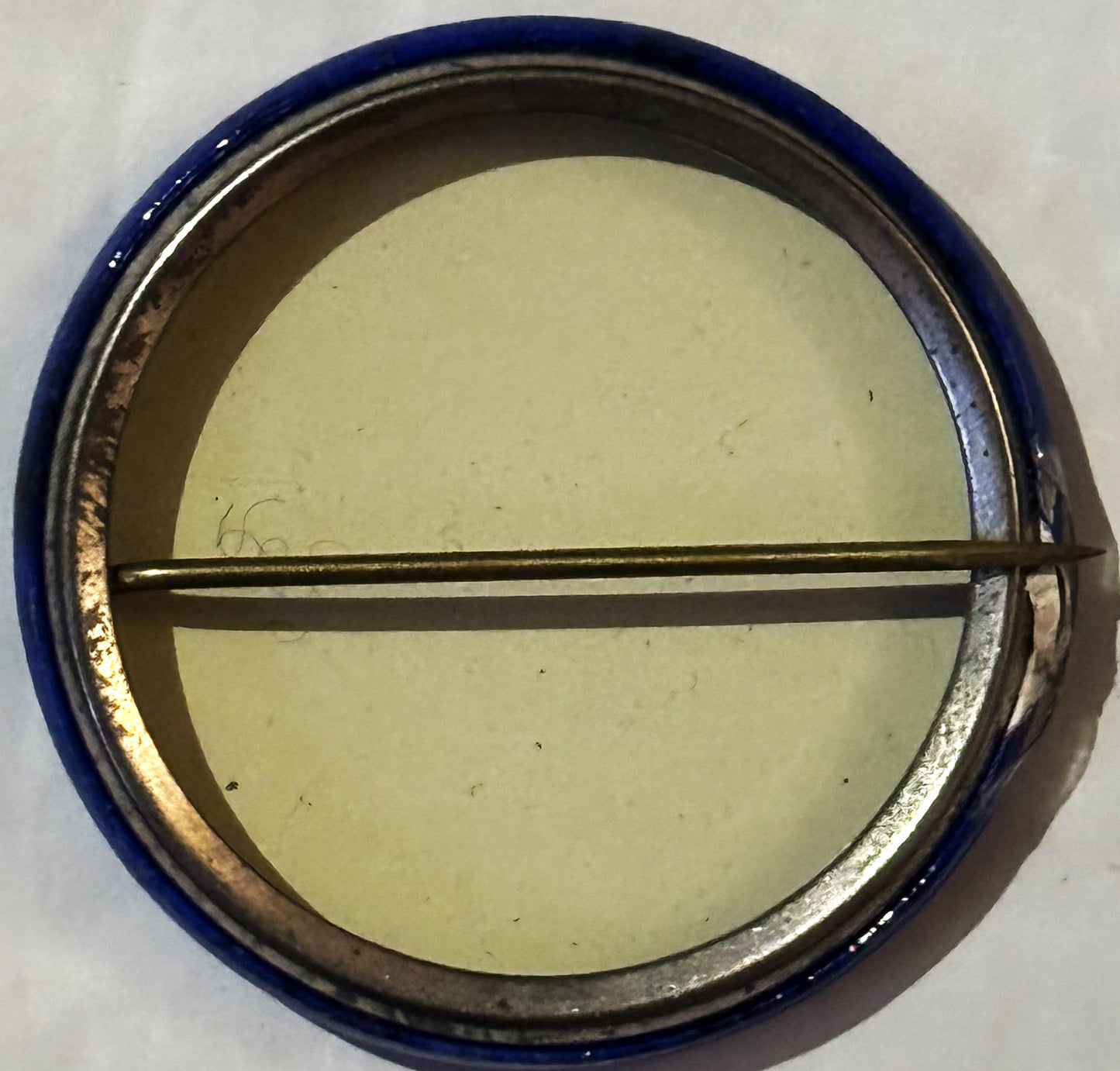 Overhead view of a vintage pinback button in a metal tin with blue rim, showing pale yellow backing and a horizontal metal cl