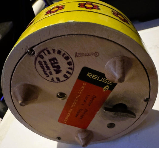 Bottom view of yellow and white vintage music box with red Reuge label and circular maker's stamp, showing mechanical details