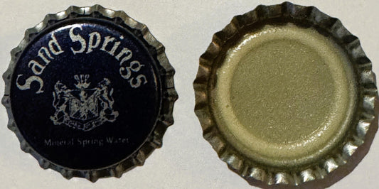 Vintage bottle cap with ridged metal edge, dark blue front showing "Sand Springs" text and coat of arms emblem, plain metalli