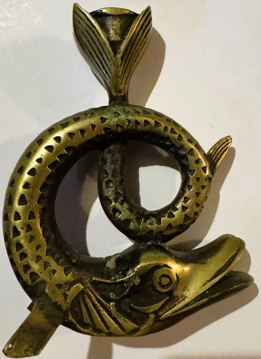 Brass fish-shaped candle holder with coiled body, detailed scales, fins, and head against white wall, casting shadow