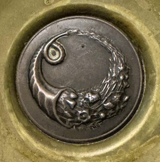 Close-up of a silver-toned circular medallion with embossed cornucopia and classical details, set in a pale brass ring dish