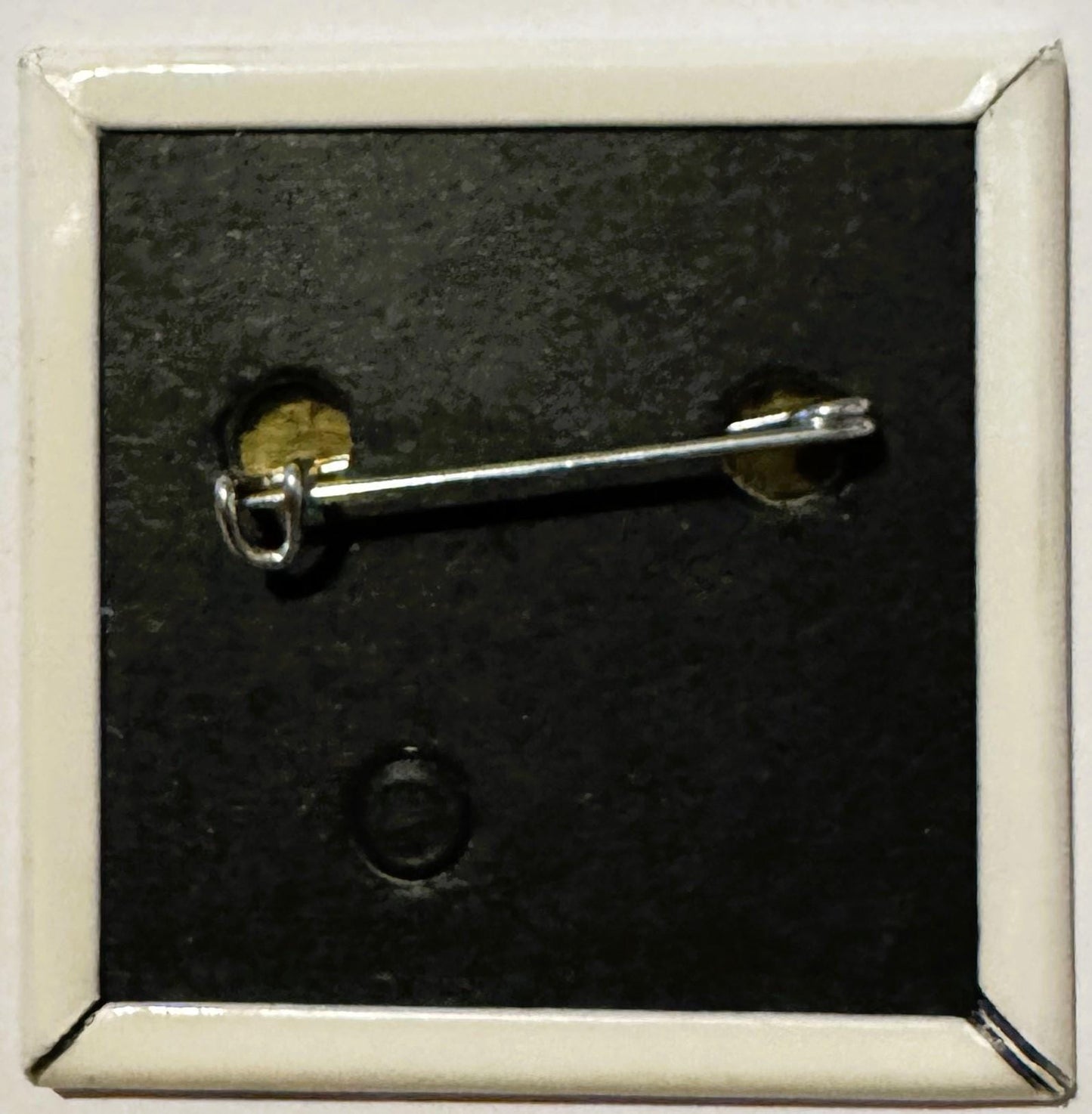 Silver pin-back button with brass fastening displayed against dark textured background in white frame, shown at angle
