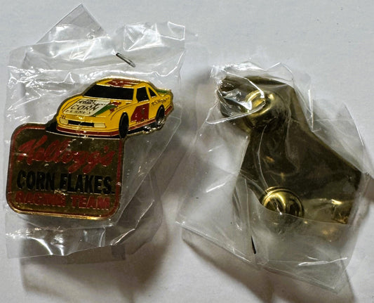 Two vintage NASCAR pins in clear plastic bags on white surface: yellow and red race car pin with "Corn Flakes" text, and bron