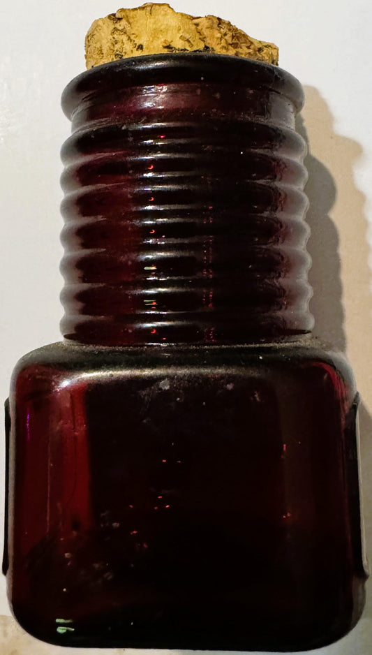 Deep ruby red glass apothecary bottle with horizontal ridged neck, cork stopper, and square base against white background