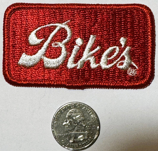 Red embroidered patch with white script "Bike's" lettering beside a quarter coin for scale