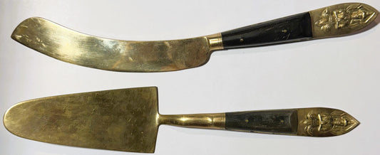 Two brass serving utensils with black handles and embossed Thai deity finials, displayed flat against white background