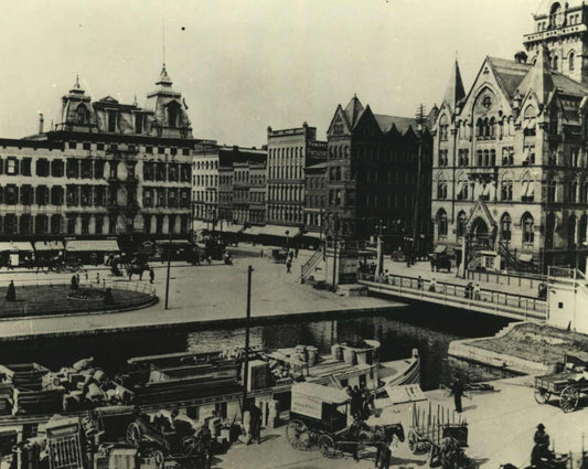 Black and white vintage photograph showing a historic waterfront cityscape with multi-story buildings, a canal, boats, horse-